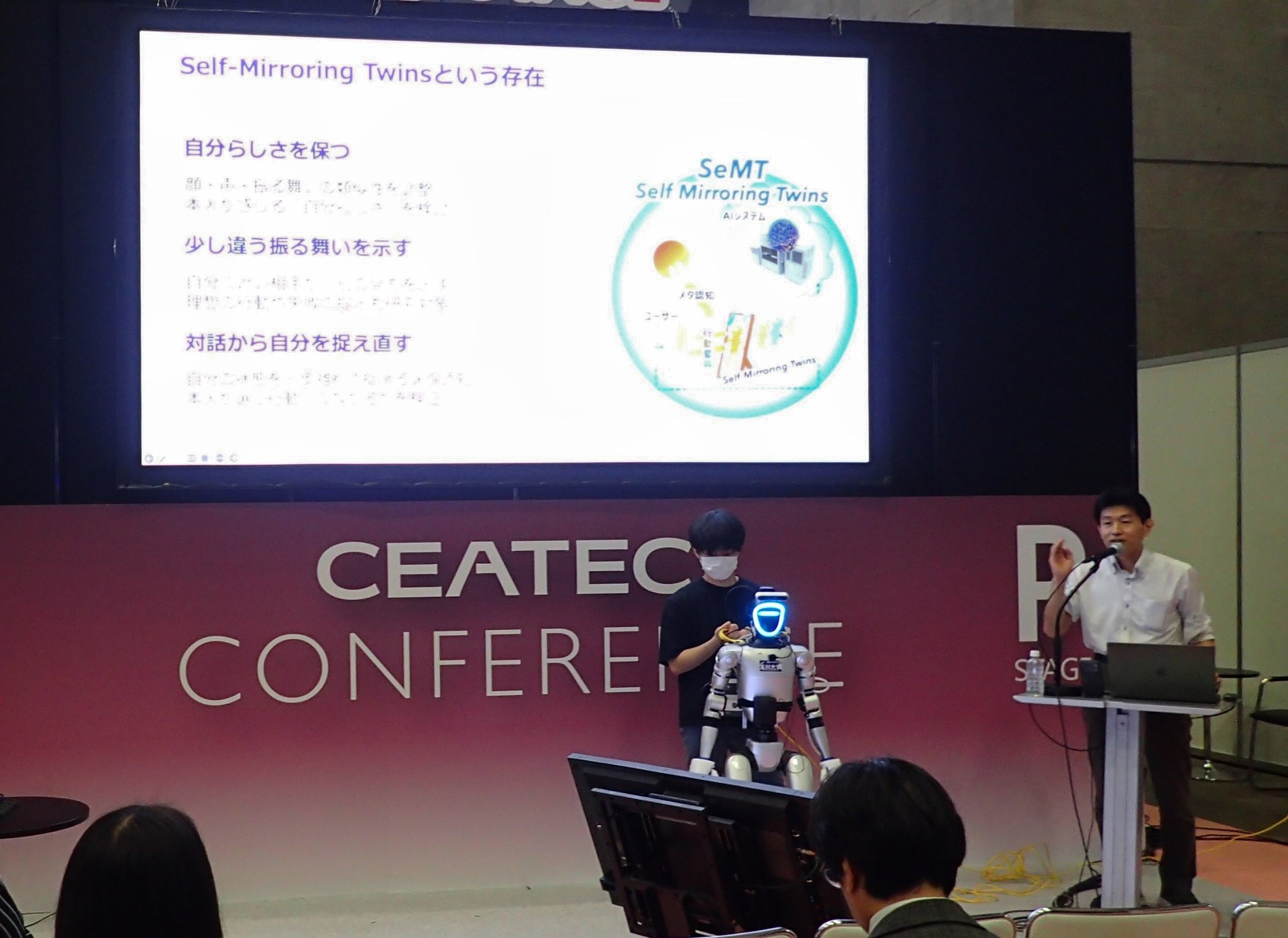 CEATEC 2026 カンファレンスのPステージで Self-Mirroring Twins について講演する稲邑哲也。スクリーンには「Self-Mirroring Twinsという存在」のスライドが映り、手前にヒューマノイドロボットが立っている。